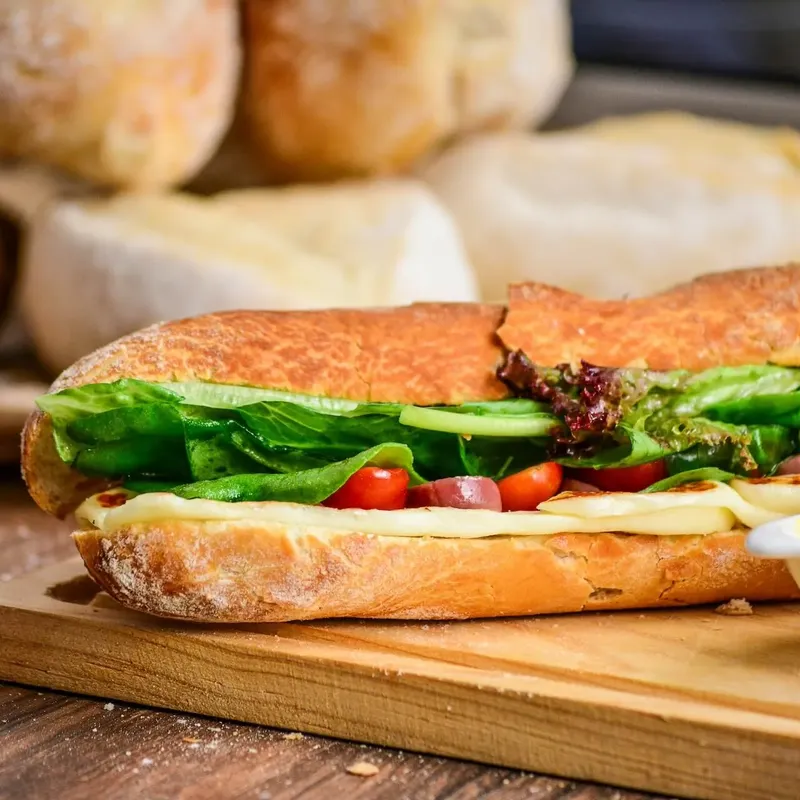 A sub with lettuce, tomato and cheese on a wooden board, bread rolls stacked behind it